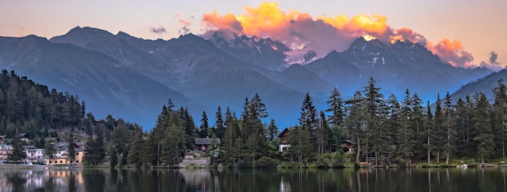 wide view of a lake with cabins and forest along the shore reflects on the surface of the water, as a beautiful sunset disappears behind a large mountainscape in the background